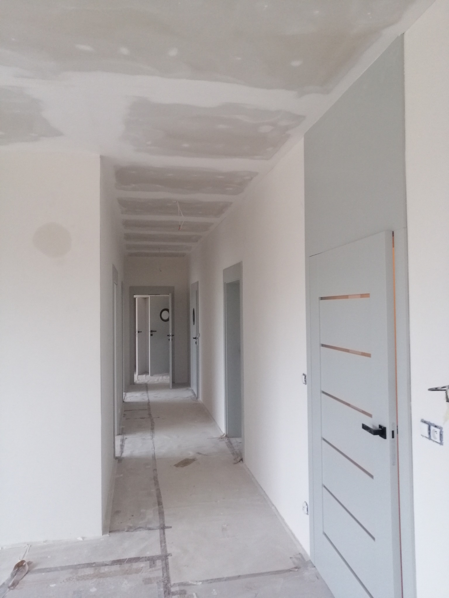 Korytarz w trakcie prac wykończeniowych: surowy sufit z widocznymi łączeniami płyt, ściany częściowo pomalowane na biało, szara ściana z nowoczesnymi drzwiami z poziomymi wstawkami, podłoga...