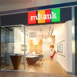 Nowoczesna aranżacja wnętrza placówki bankowej mBank z przeszklonym wejściem, drewnianą podłogą i minimalistycznym wystrojem. Widoczne biurka, krzesła i oświetlenie sufitowe.