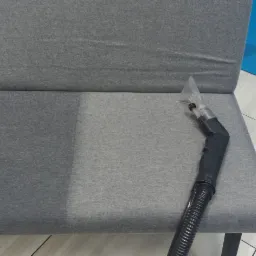 Szara kanapa z widoczną różnicą w czystości tapicerki po lewej stronie, gdzie widać ślady zabrudzeń w kształcie serca, oraz po prawej, gdzie tapicerka jest czysta i obok leży dysza odkurzacza...