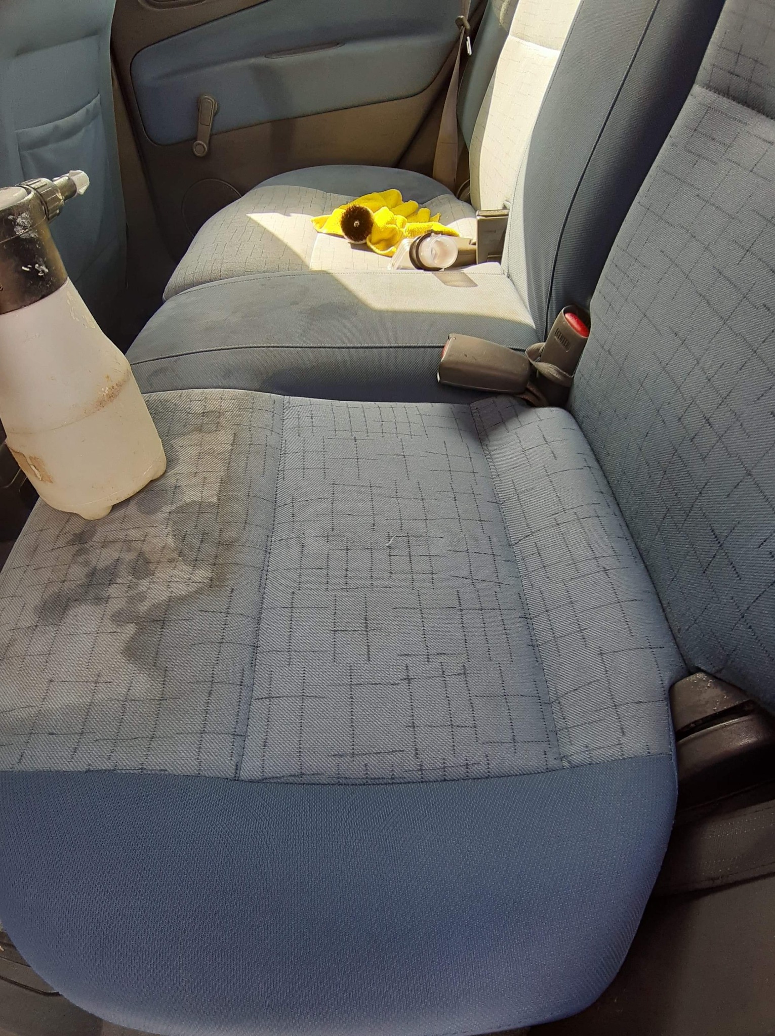 Fragment wnętrza samochodu z niebieską tapicerką fotela, na której widać plamy po czyszczeniu oraz spryskiwacz, żółta ściereczka i rękawiczka