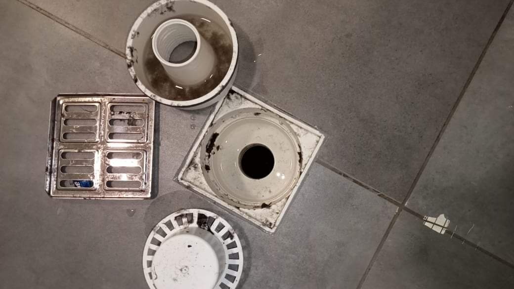 Zdemontowane, brudne elementy odpływu podłogowego, w tym kratka ściekowa, pokrywa i rura, rozłożone na szarych płytkach podłogowych.