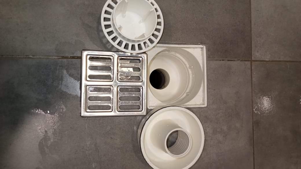 Elementy odpływu podłogowego, z metalową kratką i plastikowymi obudowami, rozłożone na szarych płytkach podłogowych, widoczne ślady wilgoci.