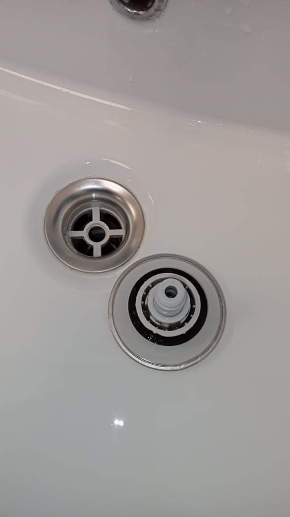 Zdemontowany, srebrny odpływ zlewu z widocznym mechanizmem wewnętrznym leżący obok zamontowanego odpływu w białej umywalce.