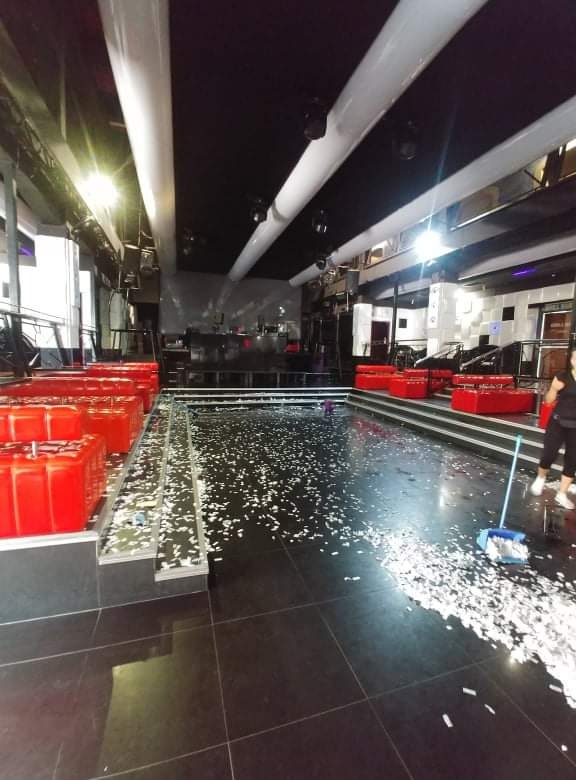 Wnętrze klubu nocnego po imprezie, podłoga pokryta konfetti, osoba sprzątająca z miotłą i szufelką, czerwone sofy w tle.