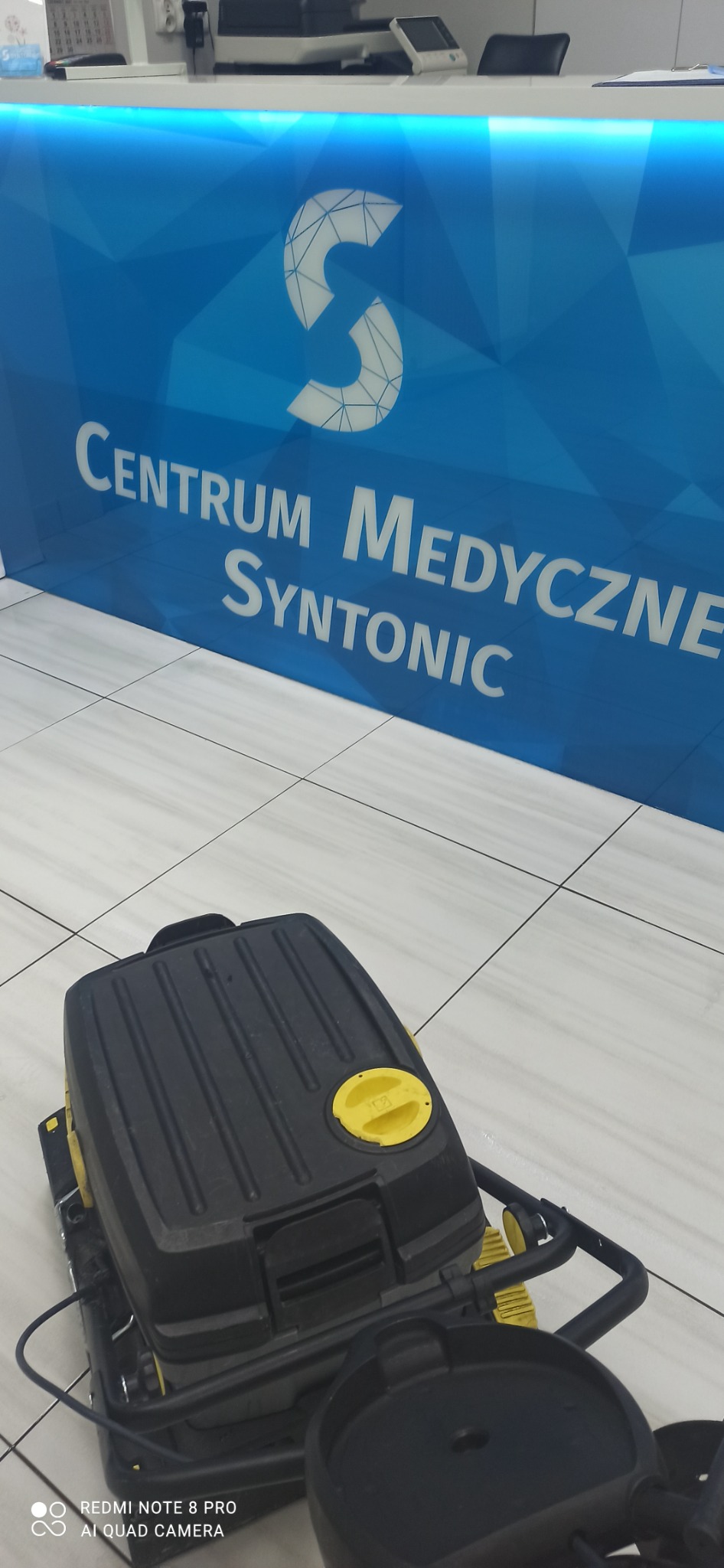 Automat szorujący do podłóg na tle lady recepcyjnej z logo Centrum Medycznego Syntonic.