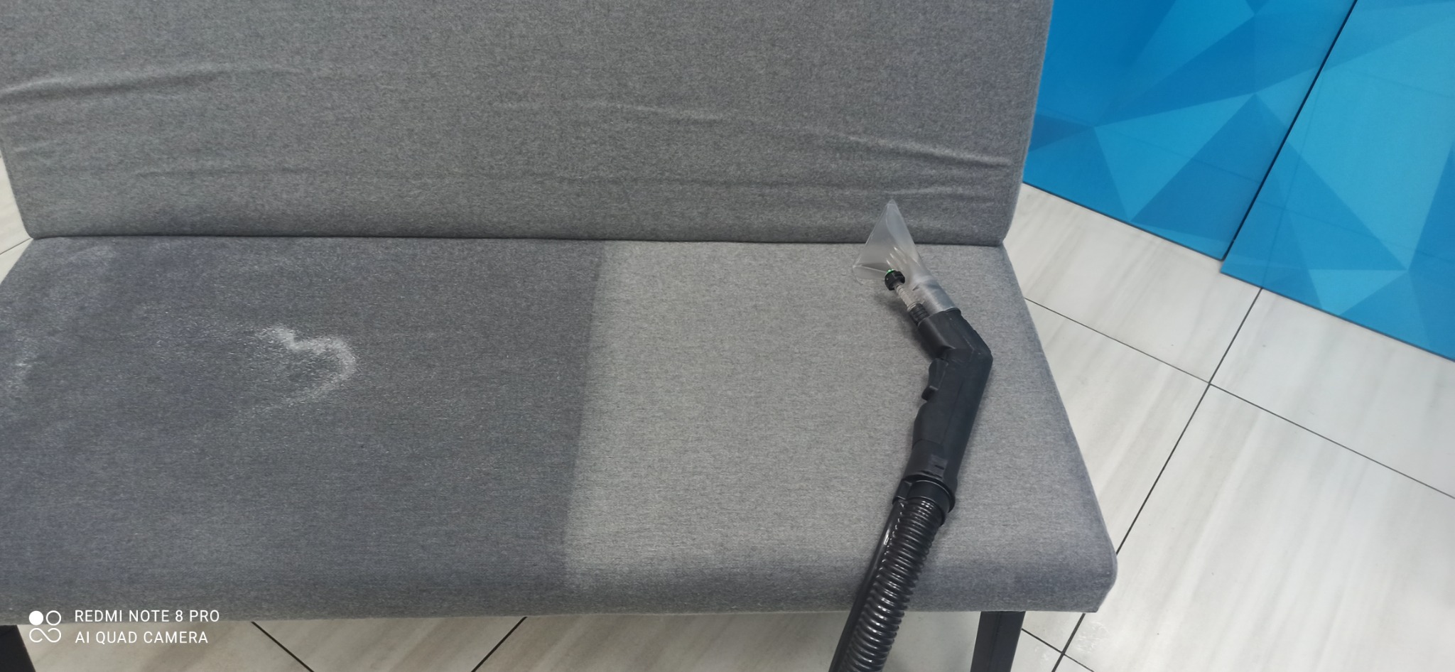 Szara kanapa z widoczną różnicą w czystości tapicerki po lewej stronie, gdzie widać ślady zabrudzeń w kształcie serca, oraz po prawej, gdzie tapicerka jest czysta i obok leży dysza odkurzacza...