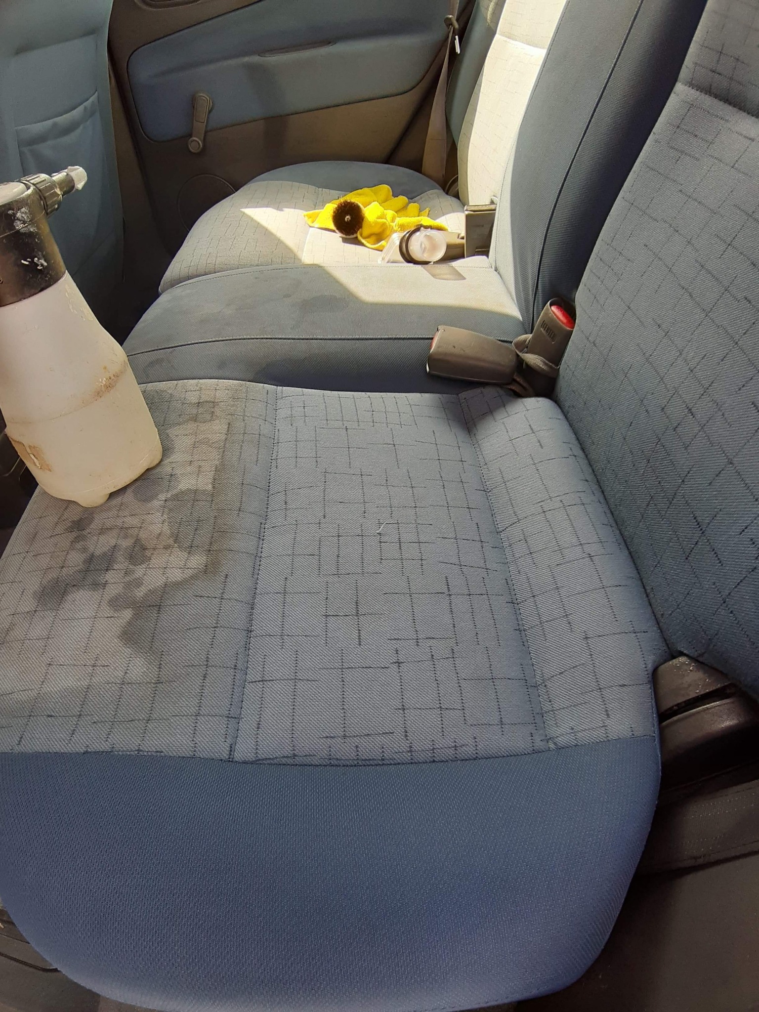 Spryskiwacz z płynem i ślady wilgoci na szarej tapicerce samochodowej z niebieskimi akcentami, obok żółta ścierka i rękawice.