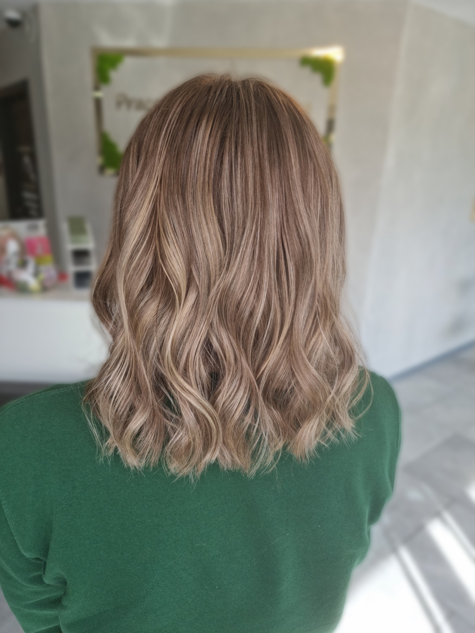 Włosy do ramion z delikatnymi falami i balejażem w odcieniach blondu i brązu, widok z tyłu, osoba w zielonej bluzce.