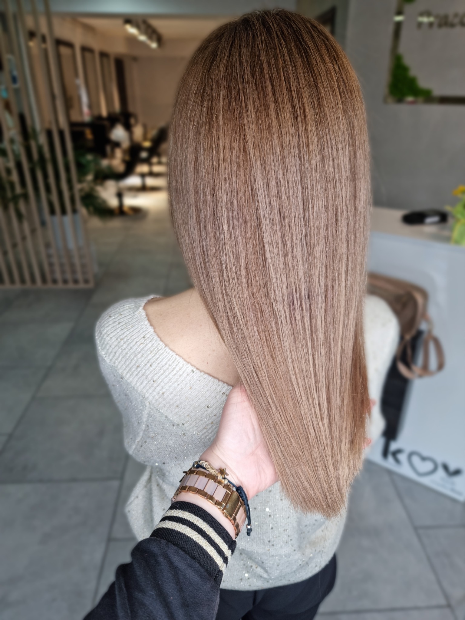 Proste, lśniące włosy w odcieniu chłodnego blondu, widok z tyłu, dłoń stylisty w paski przytrzymuje pasmo, w tle wnętrze salonu.
