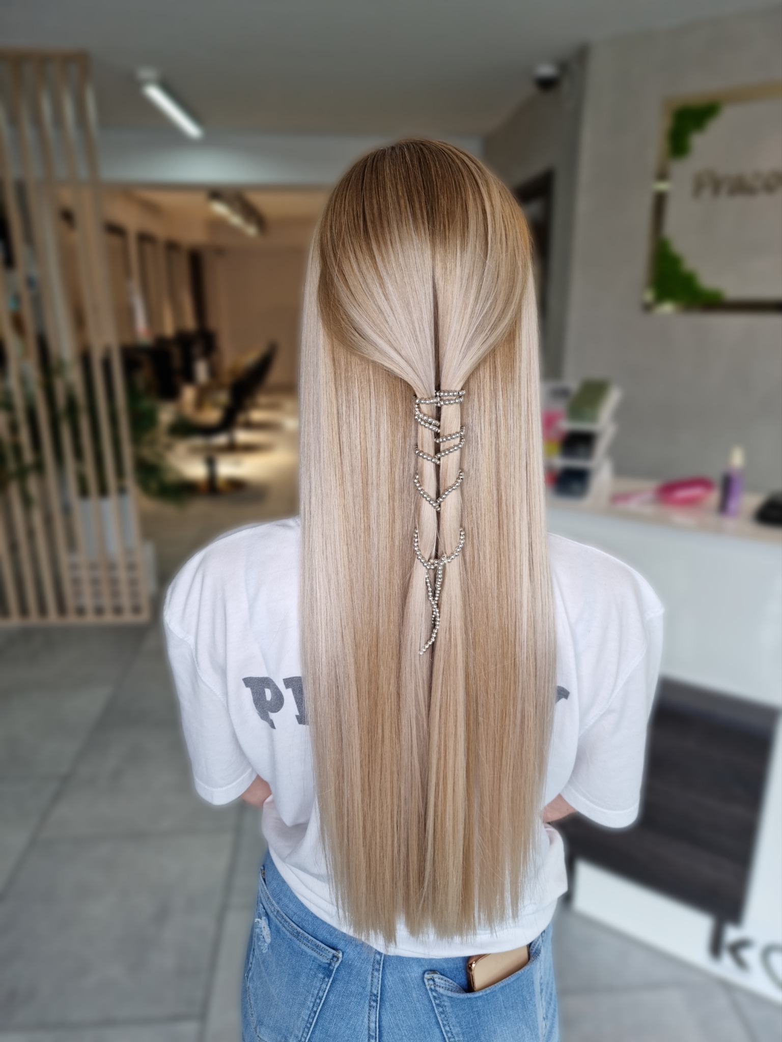 Tył głowy kobiety z długimi, prostymi, blond włosami, częściowo upiętymi ozdobną spinką z cyrkoniami, widoczna biała koszulka i dżinsy, w tle wnętrze salonu.