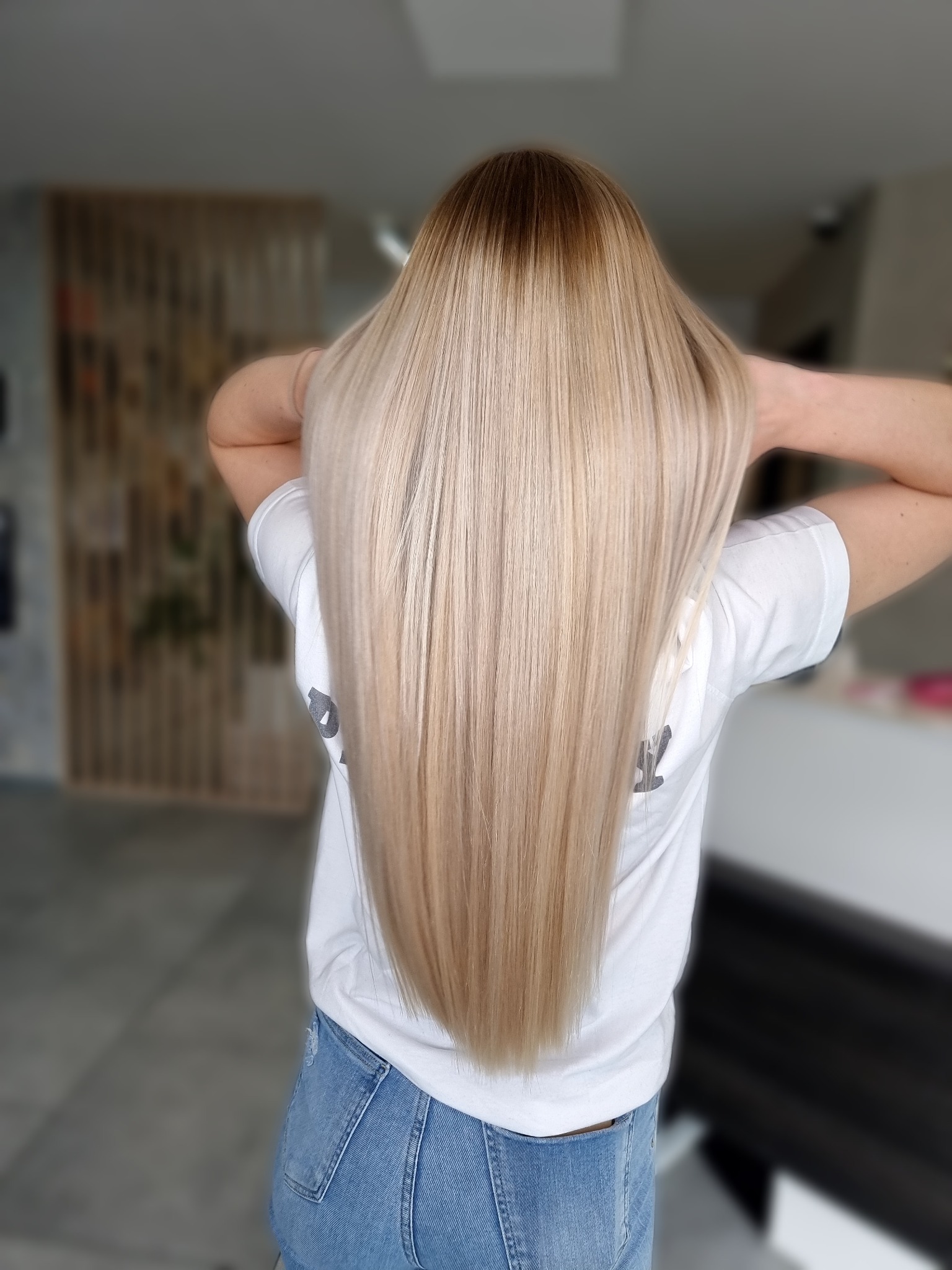 Długie, proste, blond włosy z ciemnym odrostem, widok z tyłu na tle salonu, ręce uniesione za głową.