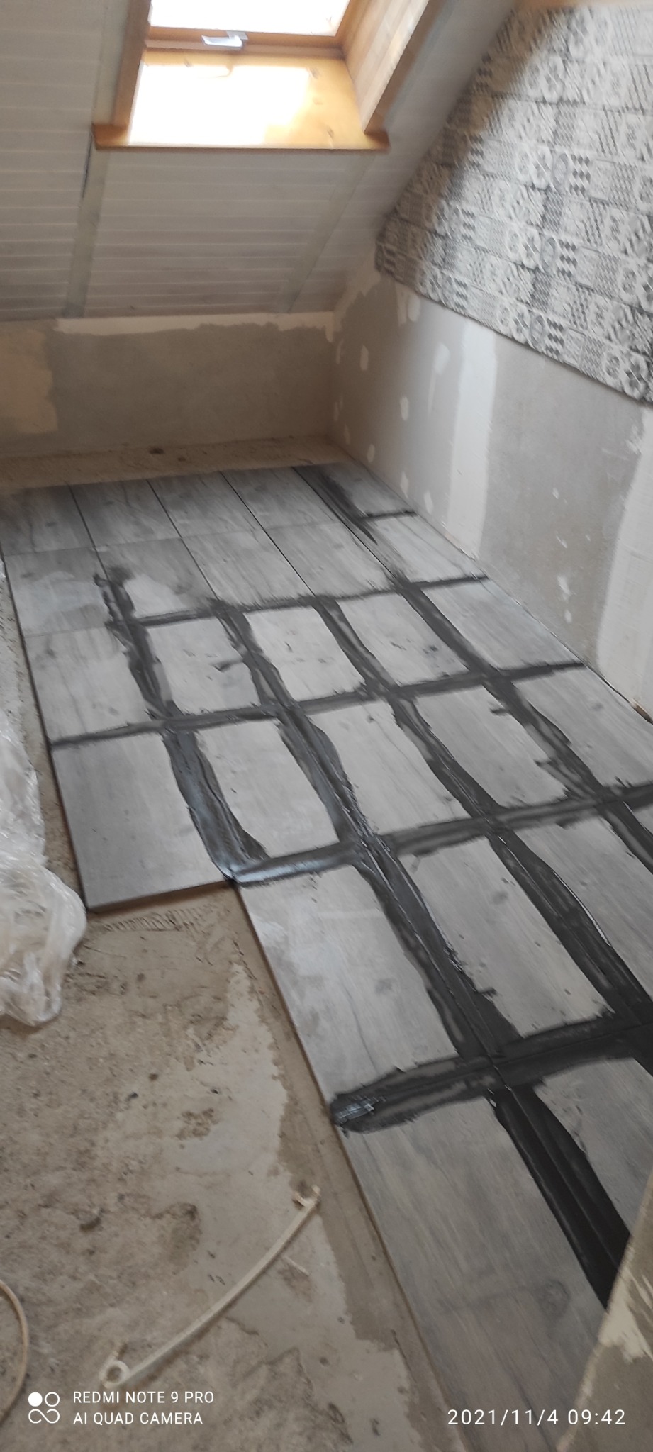 Wnętrze poddasza w trakcie układania szarej terakoty z fugą, widoczne okno dachowe, ściany przygotowane do wykończenia.