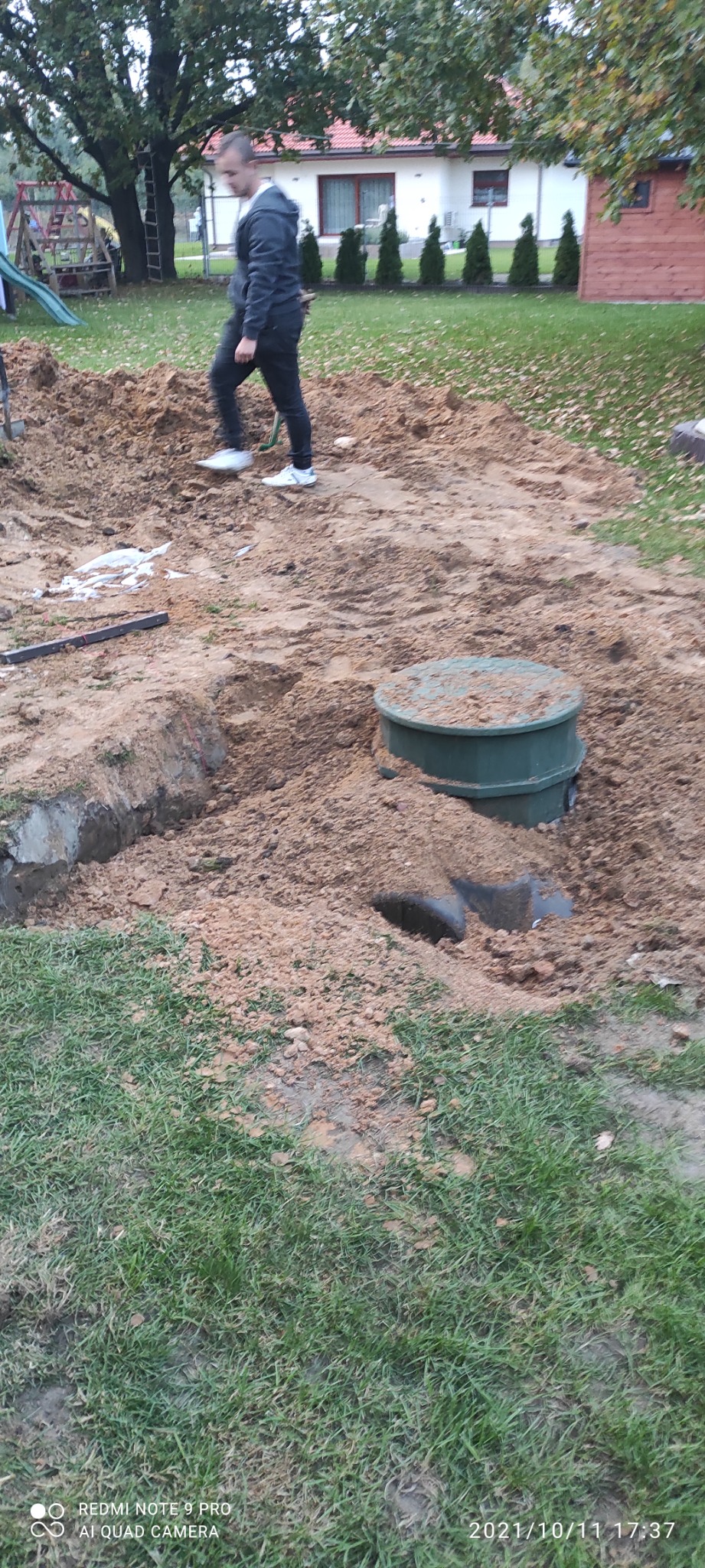 Wykopany dół z widoczną zieloną studzienką kanalizacyjną, rurą i osobą przechodzącą obok w tle, na trawniku z domem.