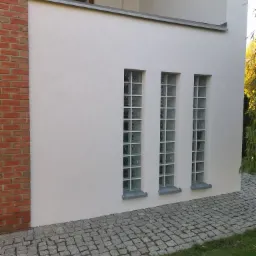 Fragment otynkowanej ściany budynku z trzema pionowymi oknami z luksferów, po lewej stronie ściana z czerwonej cegły, kostka brukowa na chodniku.