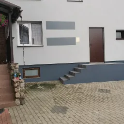 Szara elewacja domu z brązowymi drzwiami wejściowymi i dekoracyjnymi pasami w kolorze szarości, widoczne schody i bruk na podwórku.