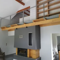 Wnętrze domu z antresolą, widoczny kominek z szarą obudową, drewniane belki stropowe, metalowa balustrada i regał na książki na antresoli, szare płytki na podłodze.