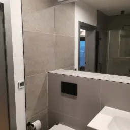 Nowoczesna łazienka z szarymi płytkami imitującymi kamień, wiszącą toaletą, białą umywalką i dużym lustrem odbijającym kabinę prysznicową.