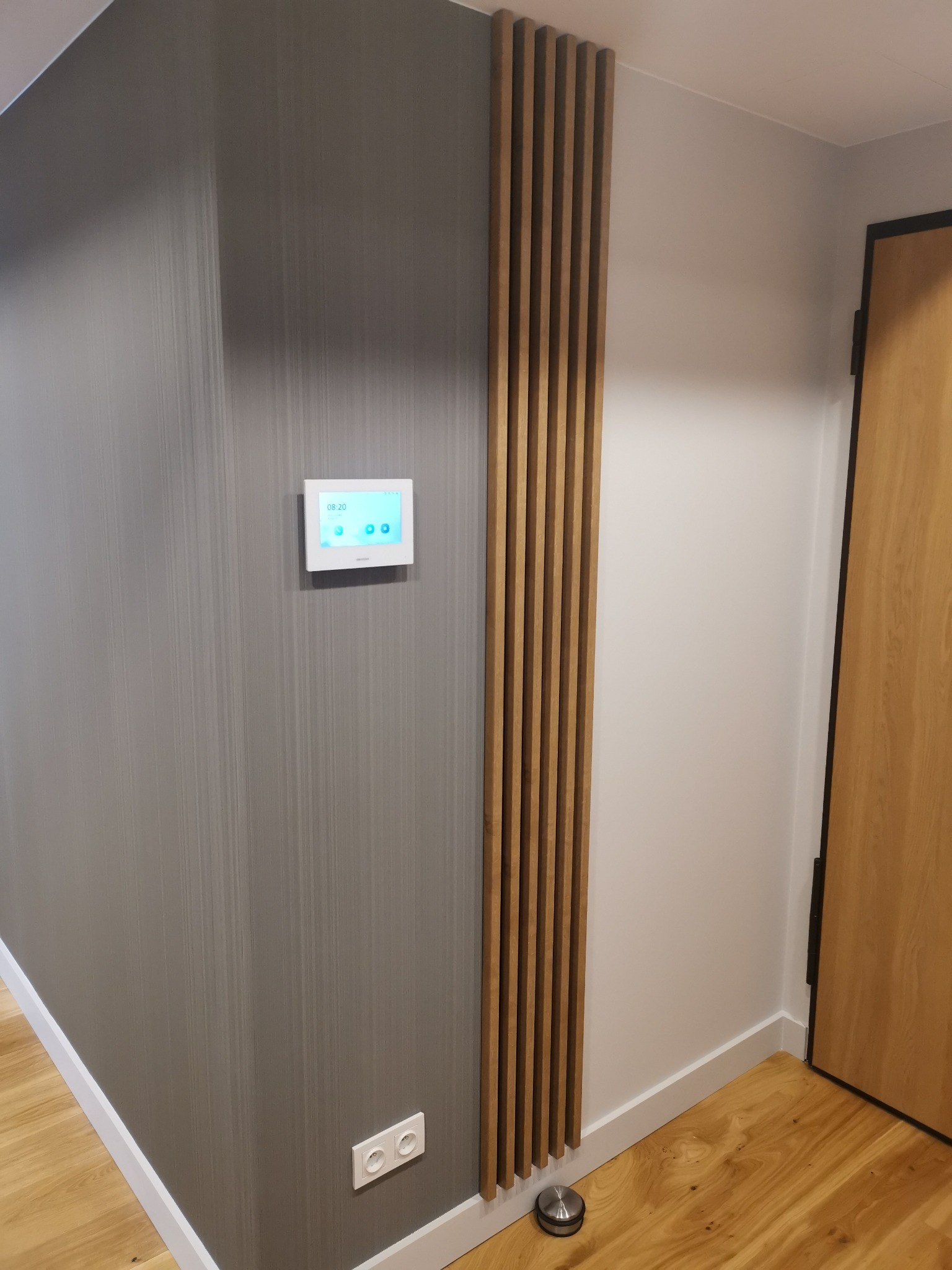 Wnętrze: szara ściana z pionowymi paskami, biały panel sterowania, drewniane listwy dekoracyjne, drewniane drzwi i jasna drewniana podłoga