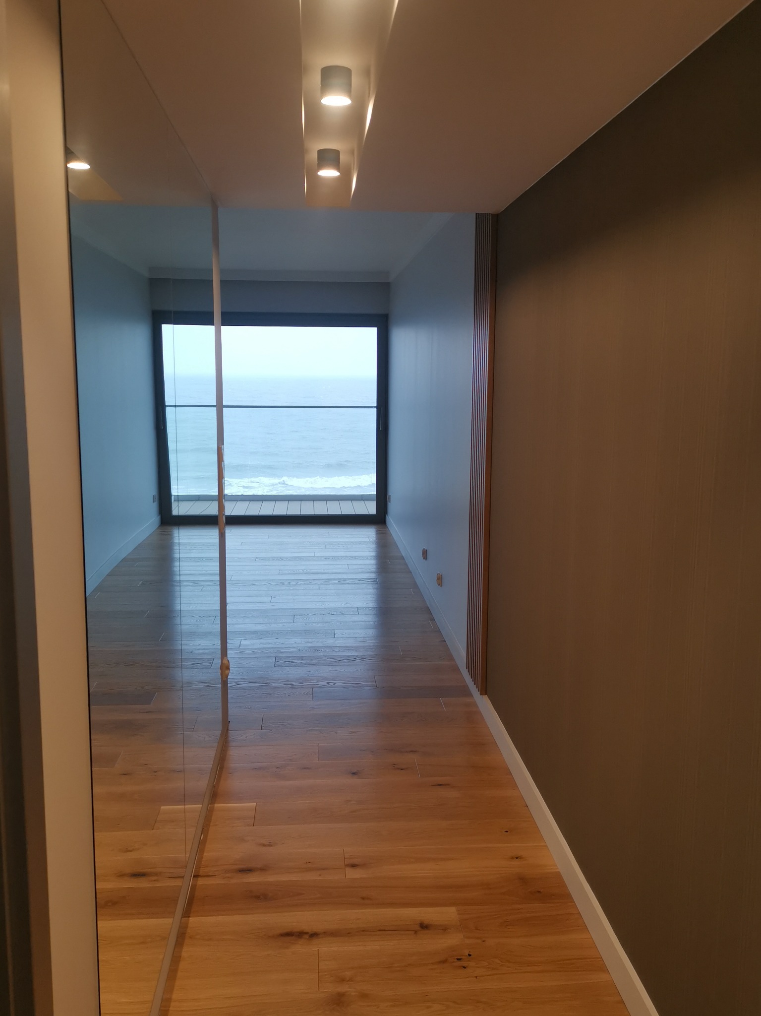 Nowoczesny korytarz z drewnianą podłogą i widokiem na morze, odbity w lustrzanej szafie, oświetlony trzema punktowymi lampami na suficie.