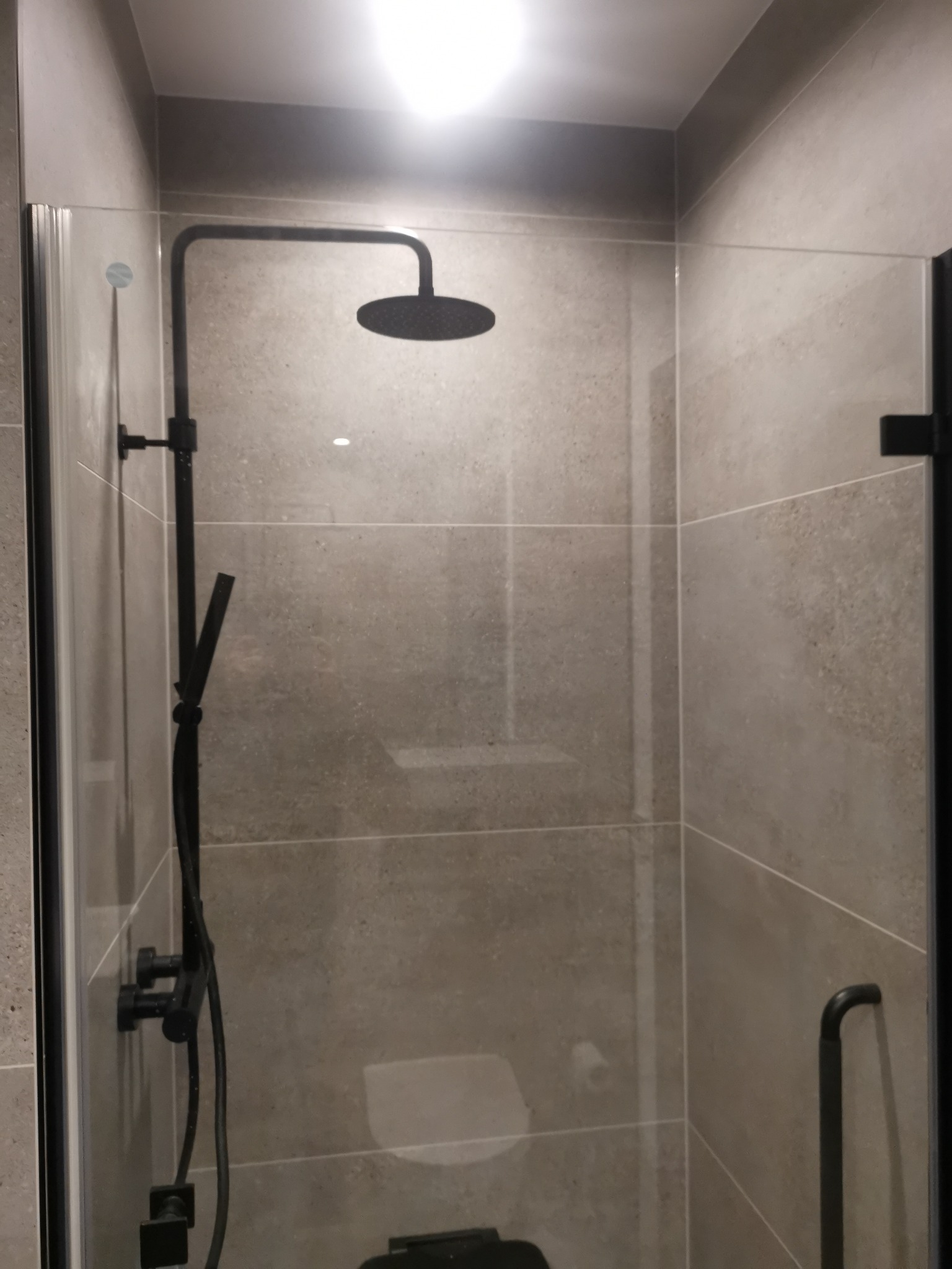 Nowoczesna kabina prysznicowa z czarną armaturą, szklane ściany z czarnymi ramami, płytki w odcieniach szarości.