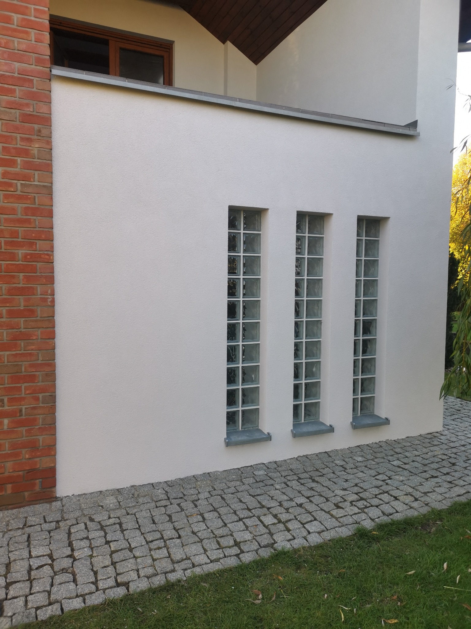 Fragment otynkowanej ściany budynku z trzema pionowymi oknami z luksferów, po lewej stronie ściana z czerwonej cegły, kostka brukowa na chodniku.