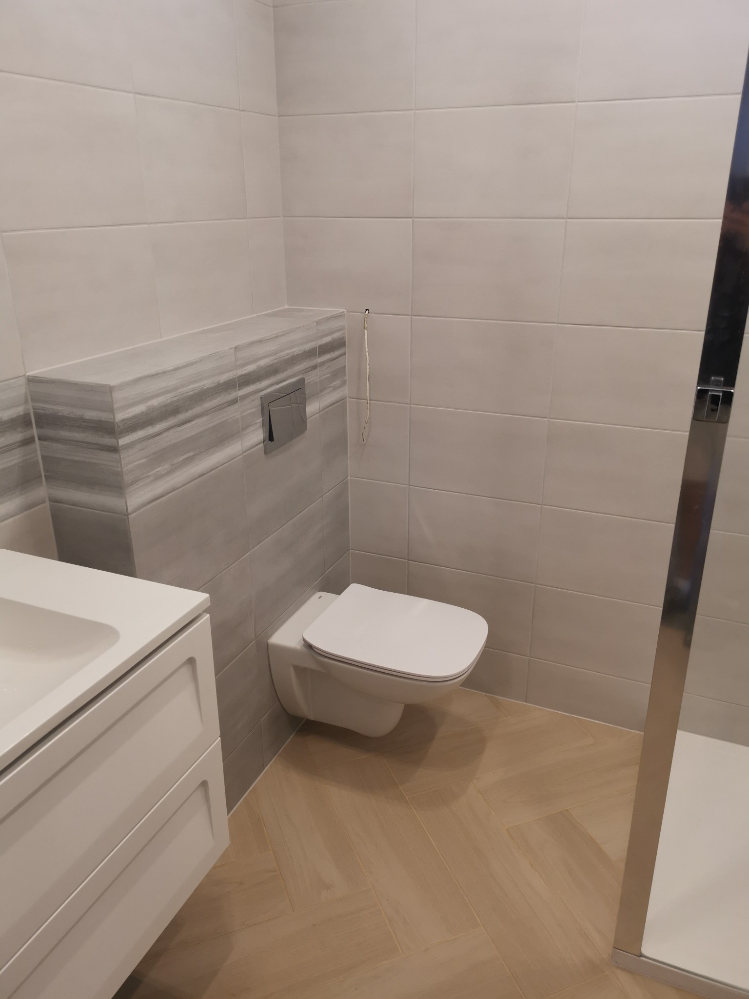 Nowoczesna łazienka w Gostyniu po renowacji: biała umywalka z szafką, podwieszany sedes z szarym panelem, jasne płytki na ścianach i podłodze ułożone w jodełkę, kabina prysznicowa z chromowanymi...