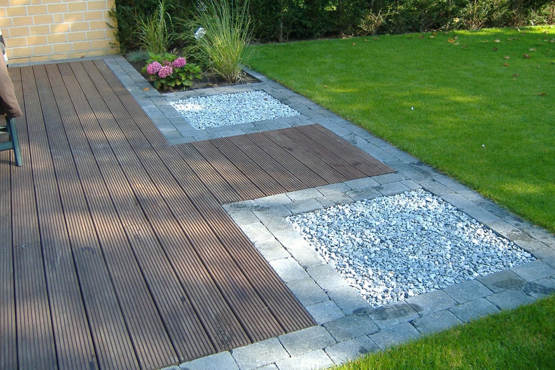 Nowoczesny taras z desek kompozytowych w kolorze ciemnego brązu, otoczony szarą kostką brukową i białym żwirem, z fragmentem trawnika i rabatą z hortensjami w tle.