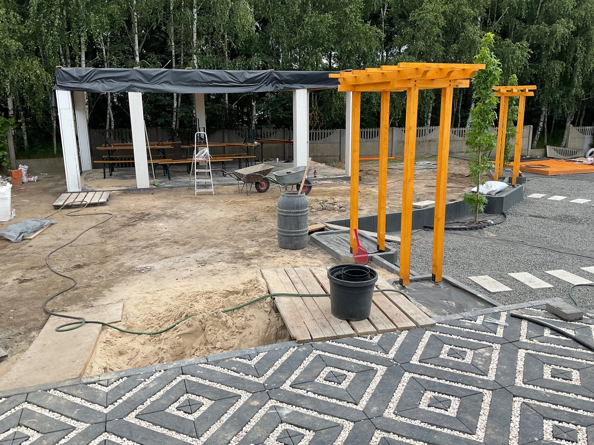 Trwają prace wykończeniowe w ogrodzie: układanie kostki brukowej o geometrycznym wzorze z białym wypełnieniem, montaż drewnianej pergoli w kolorze miodowym, budowa zadaszenia z czarną plandeką...
