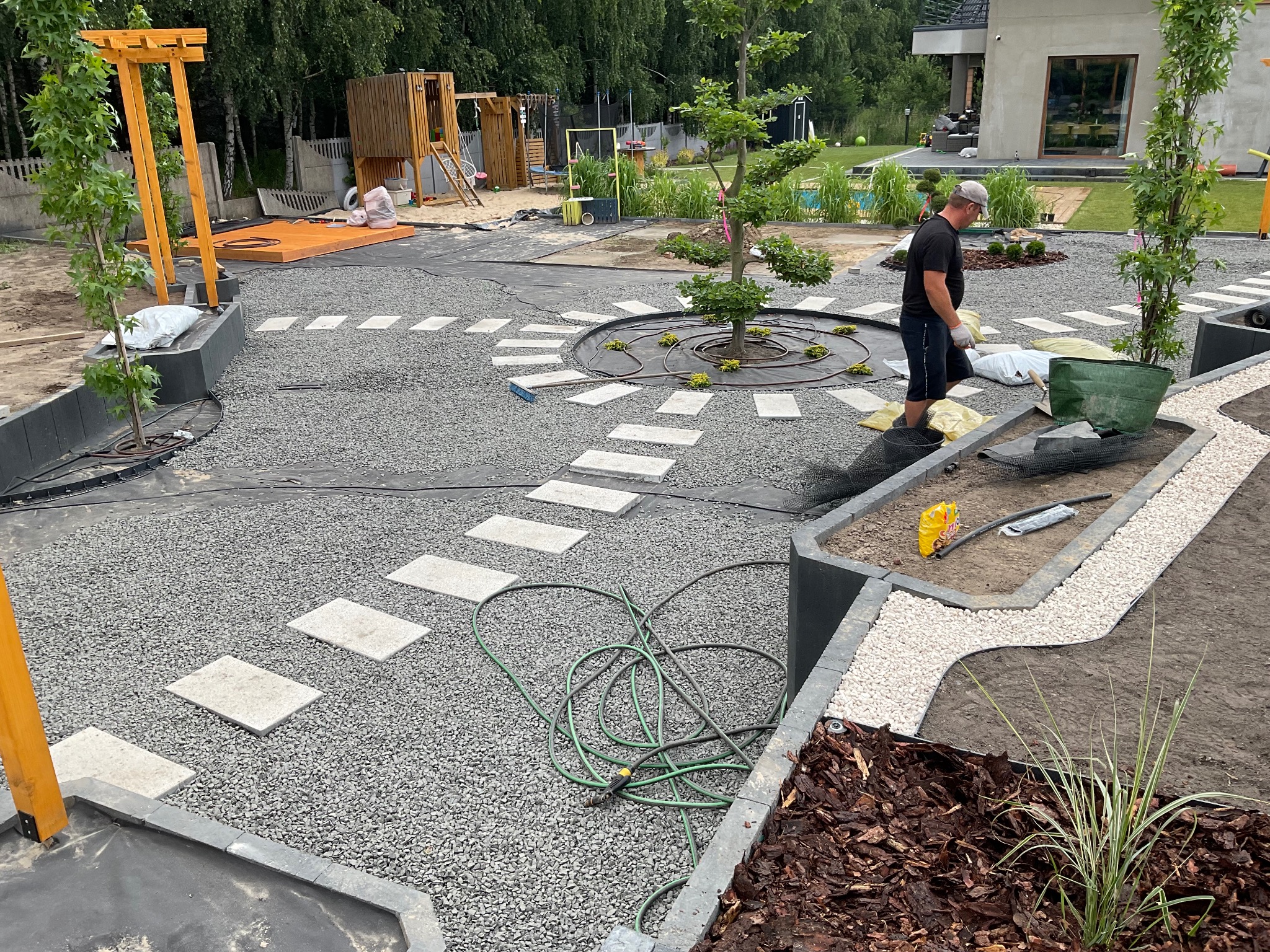 Pracownik w trakcie układania ścieżki z płyt chodnikowych na żwirowym podłożu w ogrodzie, widoczne elementy systemu nawadniania, przygotowane rośliny i kora ozdobna.