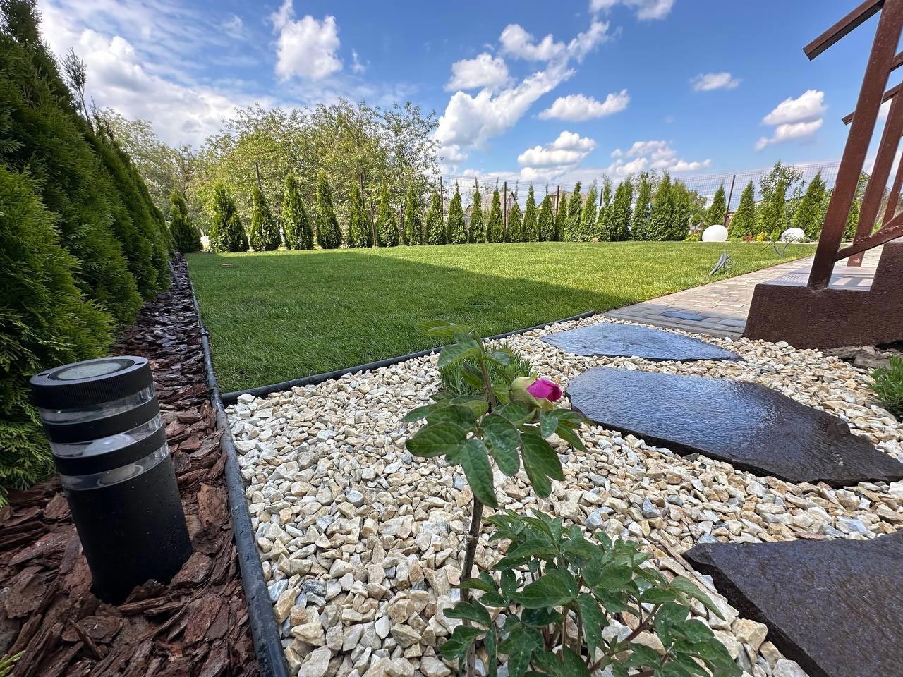 Uporządkowany ogród z równo przystrzyżonym trawnikiem, ozdobnymi kamieniami, ścieżką z nieregularnych płyt kamiennych i niskimi tujami wzdłuż ogrodzenia, oświetlony lampą ogrodową.