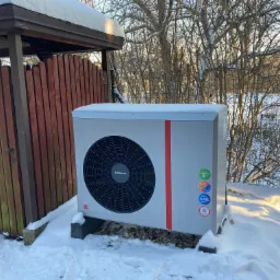 Biała pompa ciepła marki Kolton, stojąca na betonowych wspornikach w zasypanym śniegiem ogrodzie, z widoczną drewnianą wiatą i drzewami w tle.