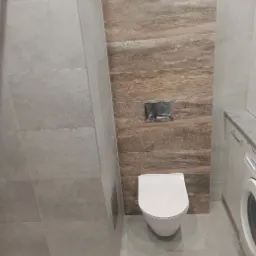 Nowoczesna łazienka z płytkami imitującymi drewno na ścianie, wisząca toaleta i pralka w zabudowie.