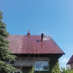 Pracownik w kasku, z uprzężą, czyści czerwoną, metalową dachówkę domu jednorodzinnego za pomocą myjki ciśnieniowej; widoczne plamy czystej dachówki i węże doprowadzające wodę.