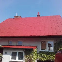Dom z nowym, intensywnie czerwonym dachem z blachy trapezowej, obrośnięty bluszczem, widoczne kominy i numer domu '42'.