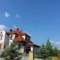 Dwupiętrowy dom z czerwoną dachówką, z pracownikiem na dachu i drabinami opartymi o elewację, widziany zza ozdobnego ogrodzenia.