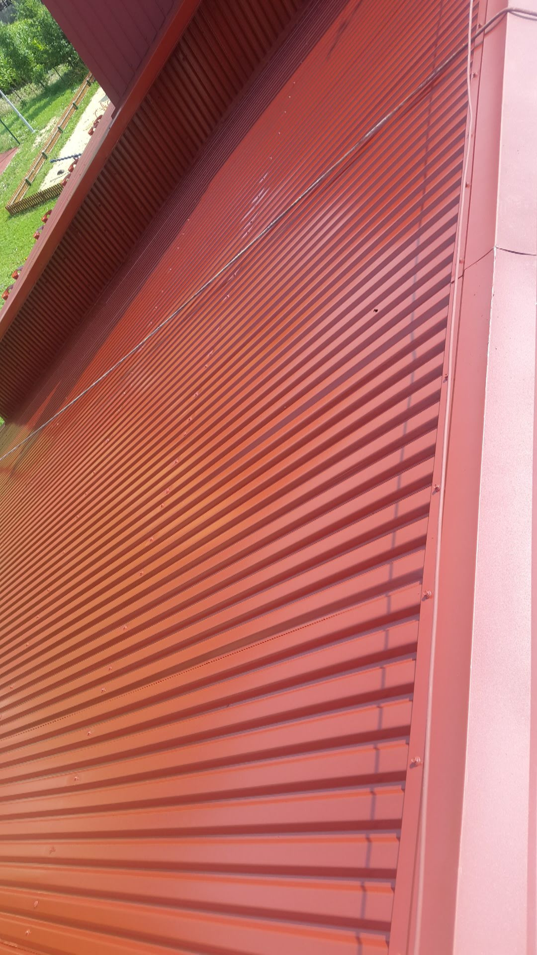Czerwona blacha trapezowa elewacyjna, widok z dołu pod kątem, z widocznymi mocowaniami i fragmentem konstrukcji budynku.