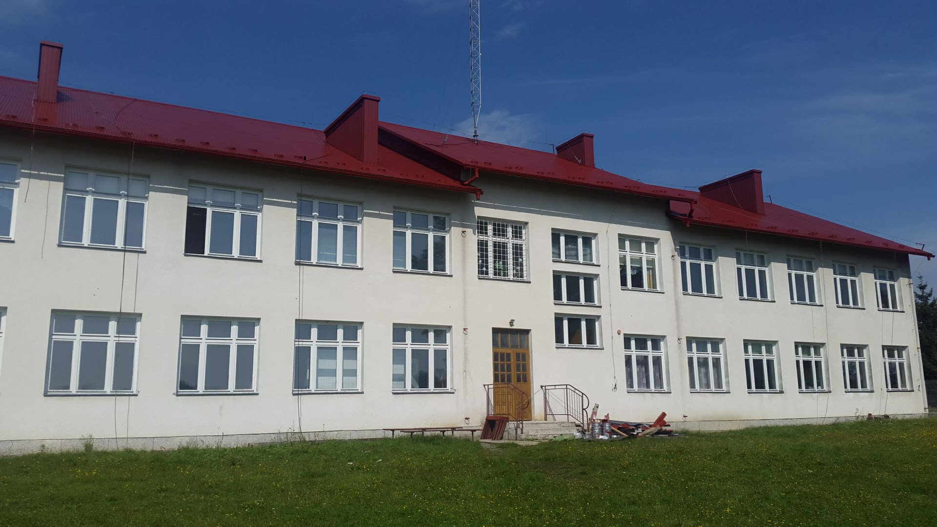 Dwupiętrowy budynek z nową, czerwoną blachodachówką, pomalowaną elewacją w odcieniach bieli i licznymi, symetrycznie rozmieszczonymi oknami z białymi ramami; na pierwszym planie trawnik z żółtymi...