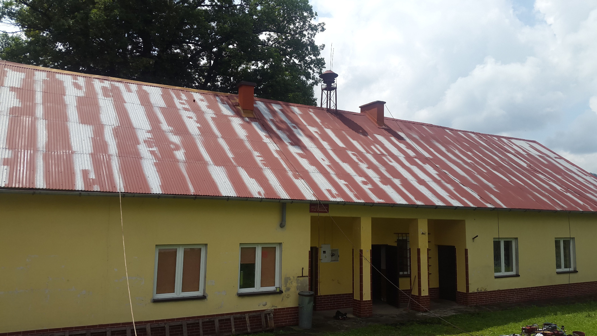 Żółty budynek z czerwonym, częściowo pomalowanym dachem z blachy falistej, widoczne ślady starej farby, kominy, maszt alarmowy na dachu, okna z białymi ramami, drabina oparta o ścianę, trawnik...