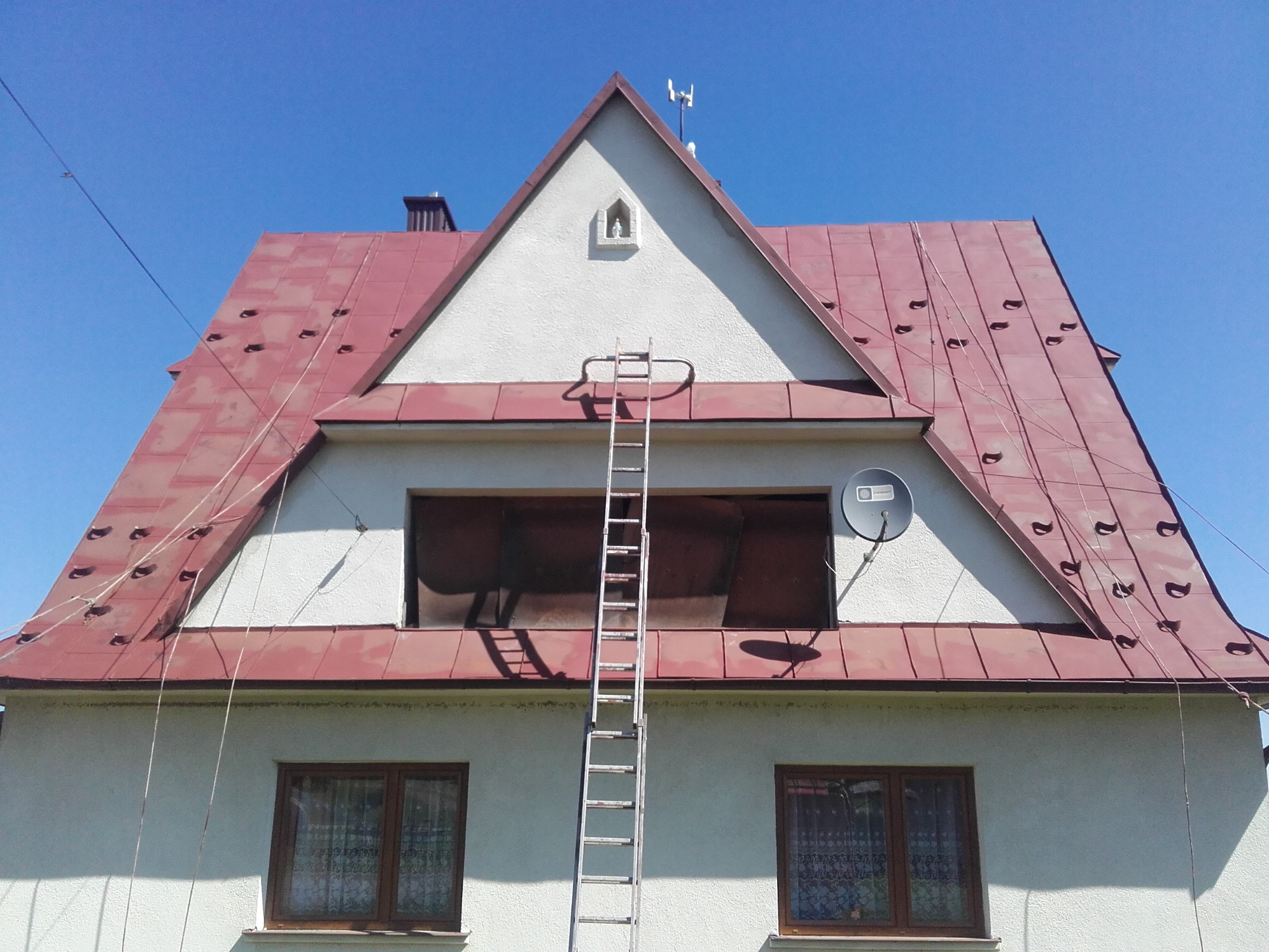 Dwupiętrowy dom z czerwoną blachodachówką, aluminiowa drabina oparta o okno na poddaszu, antena satelitarna po prawej stronie, figurka w niszy na szczycie.