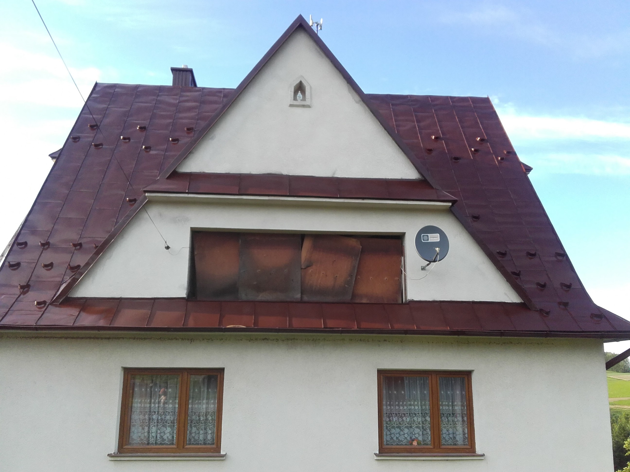 Dwuspadowy dach z blachy w kolorze ciemnej czerwieni, z widocznymi elementami wentylacyjnymi, na tle białej elewacji domu z dwoma oknami i zamurowanym otworem okiennym na poddaszu.
