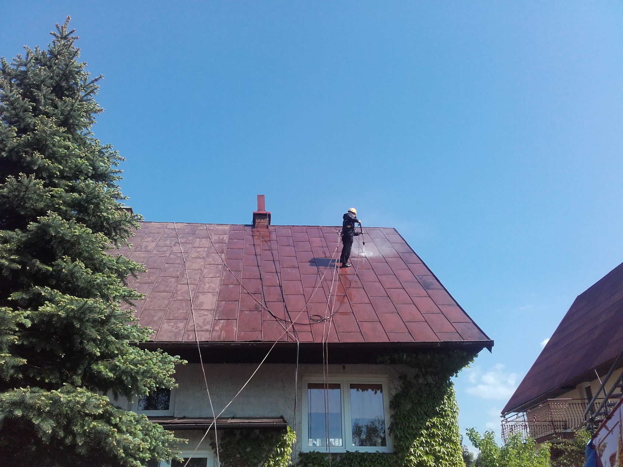 Pracownik w kasku, z uprzężą, czyści czerwoną, metalową dachówkę domu jednorodzinnego za pomocą myjki ciśnieniowej; widoczne plamy czystej dachówki i węże doprowadzające wodę.
