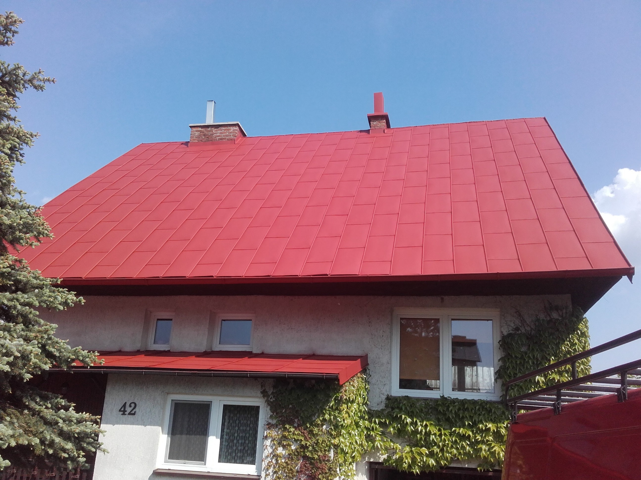 Dom z nowym, intensywnie czerwonym dachem z blachy trapezowej, obrośnięty bluszczem, widoczne kominy i numer domu '42'.