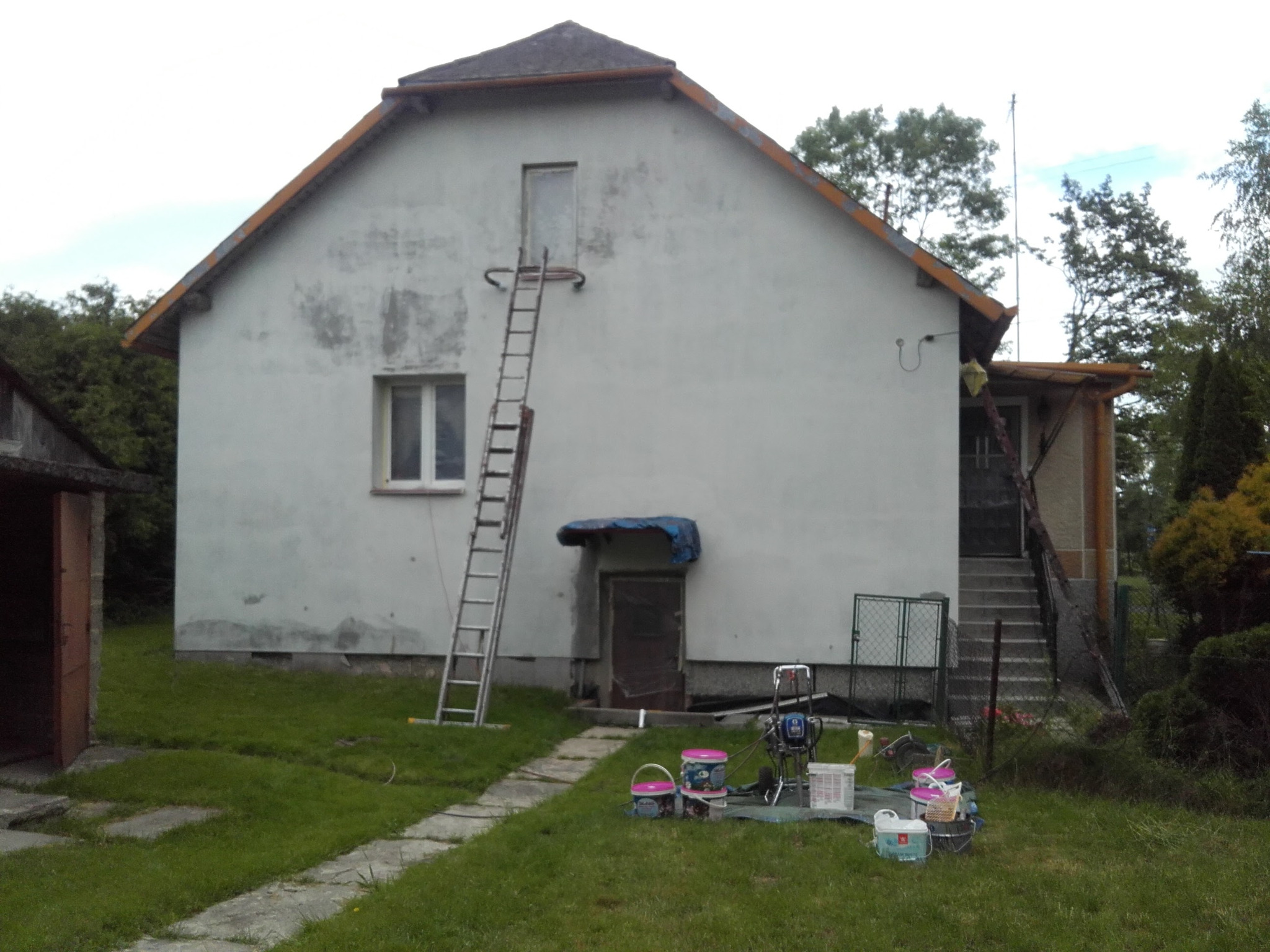 Przygotowania do malowania elewacji domu jednorodzinnego: drabina oparta o ścianę, wiaderka z farbą i agregat malarski na trawniku, widoczne ślady wcześniejszych prac malarskich na elewacji.