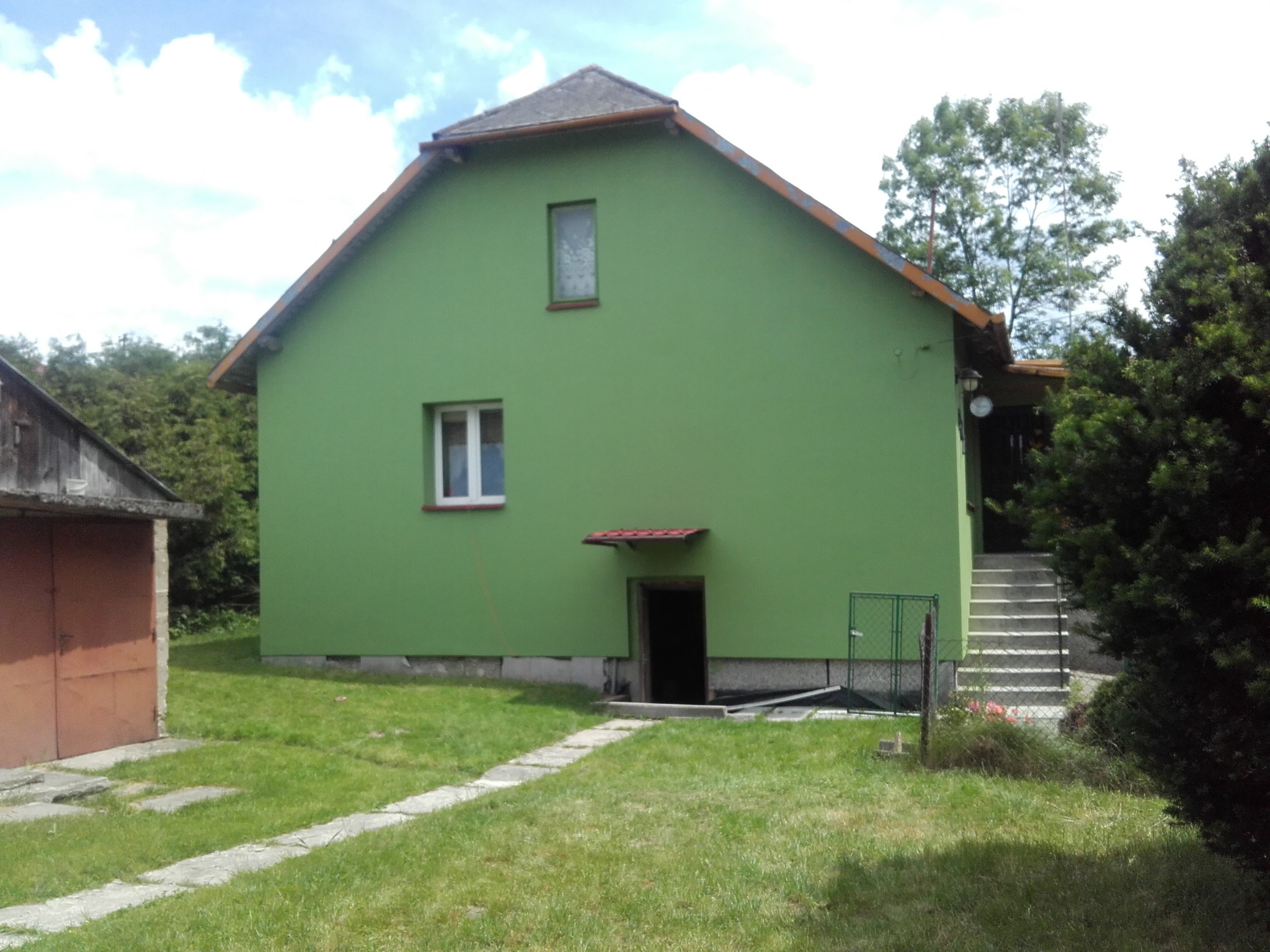 Świeżo pomalowany na zielono dom jednorodzinny z szarą podmurówką i małym oknem nad wejściem do piwnicy, widoczny z boku, z trawnikiem i ścieżką z płyt.