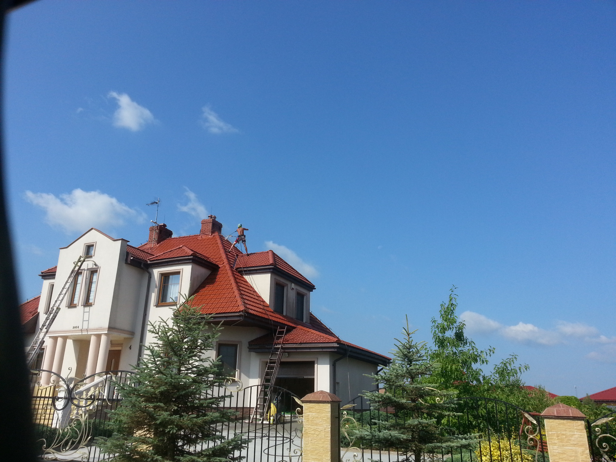 Dwupiętrowy dom z czerwoną dachówką, z pracownikiem na dachu i drabinami opartymi o elewację, widziany zza ozdobnego ogrodzenia.