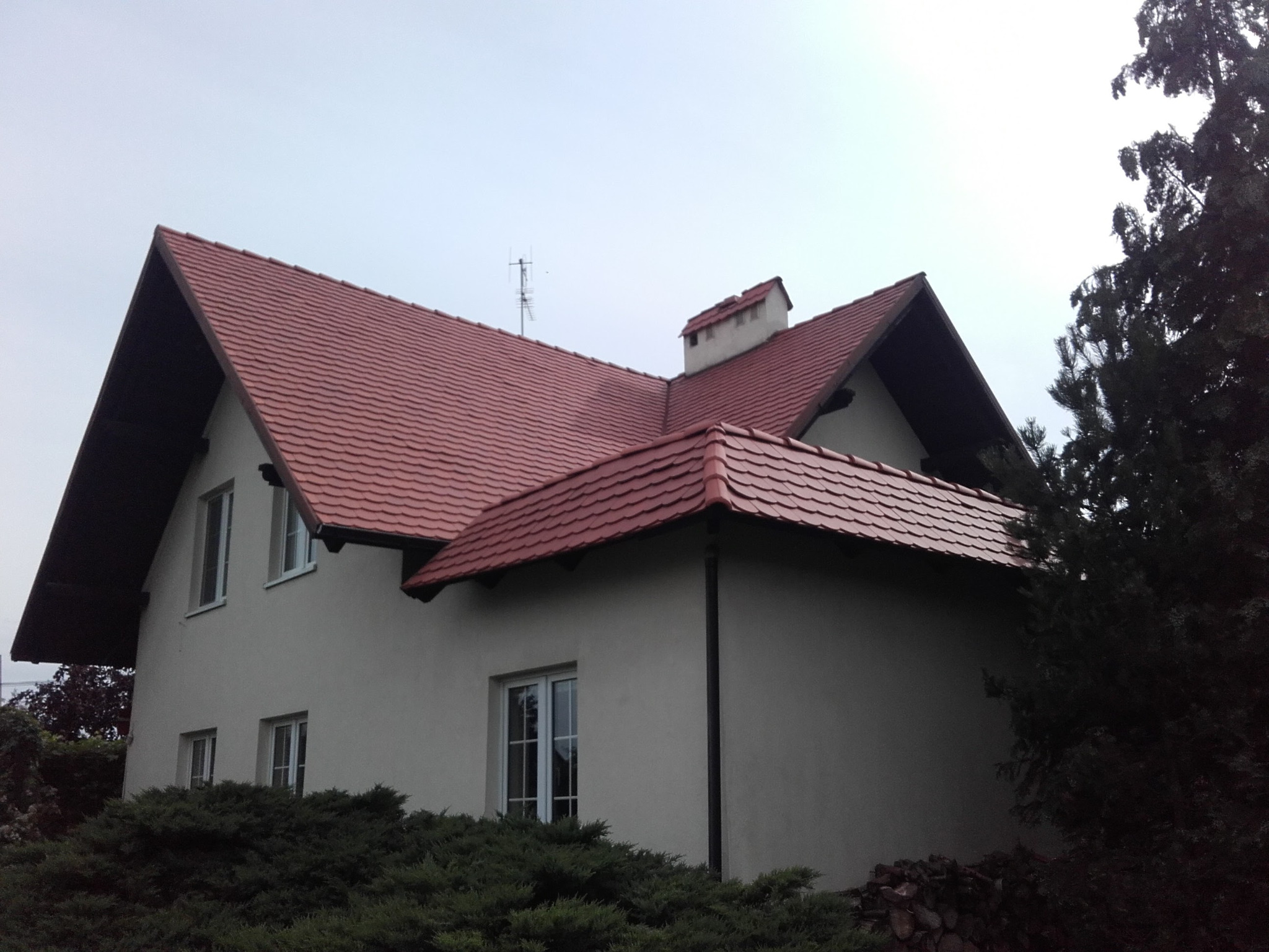 Dwupiętrowy dom z jasną elewacją i czerwoną dachówką, widoczny z niskiej perspektywy w pochmurny dzień, z anteną na dachu i bujną zielenią w ogrodzie.