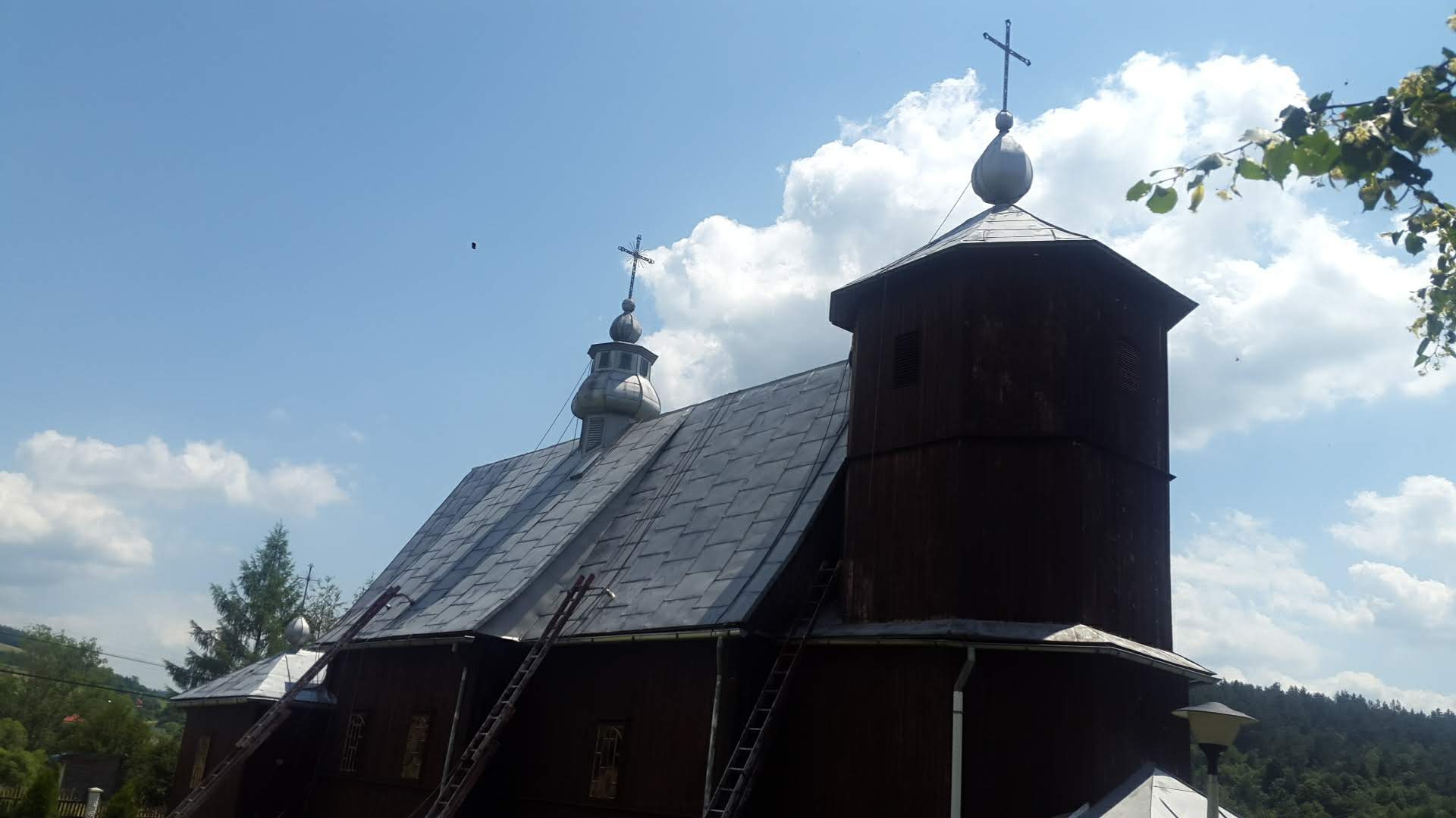 Drewniany kościół z szarą dachówką i dwoma drabinkami opartymi o elewację, krzyże na wieżyczkach, błękitne niebo z białymi obłokami w tle.