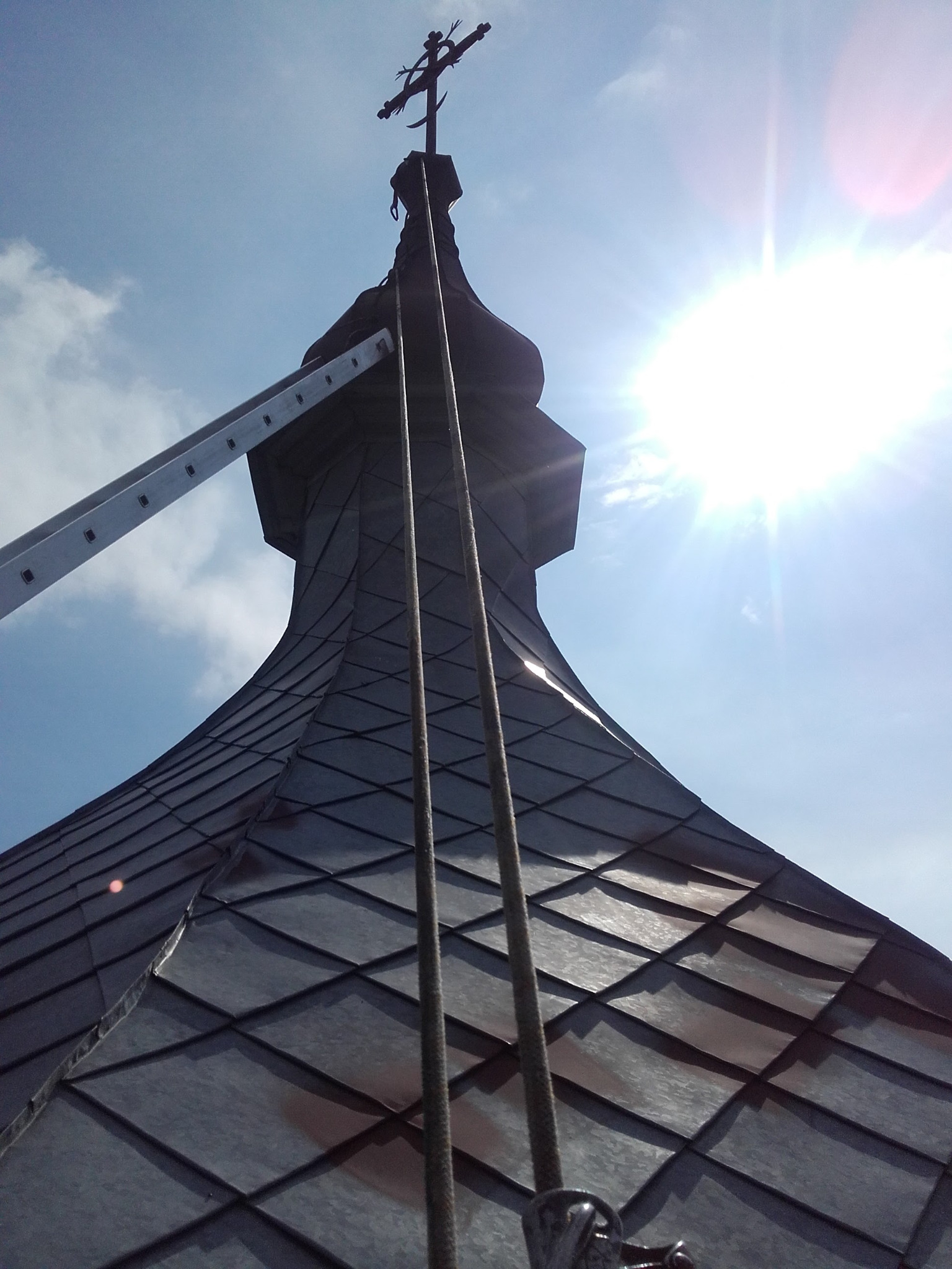 Metalowa kopuła wieży kościelnej z krzyżem na szczycie, widoczna drabina przystawiona do boku, jasne słońce na błękitnym niebie.