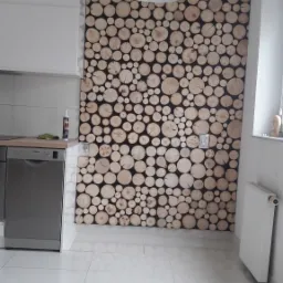 Ściana w kuchni we Wrocławiu wykończona dekoracyjnymi plastrami drewna, tworzącymi unikalną mozaikę. Białe szafki kuchenne i podłoga z jasnych płytek podkreślają nowoczesny charakter wnętrza.