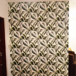 Ściana pokryta tapetą we wzór dużych, zielono-szarych liści, widoczny fragment brązowych drzwi i białego telefonu na ścianie.
